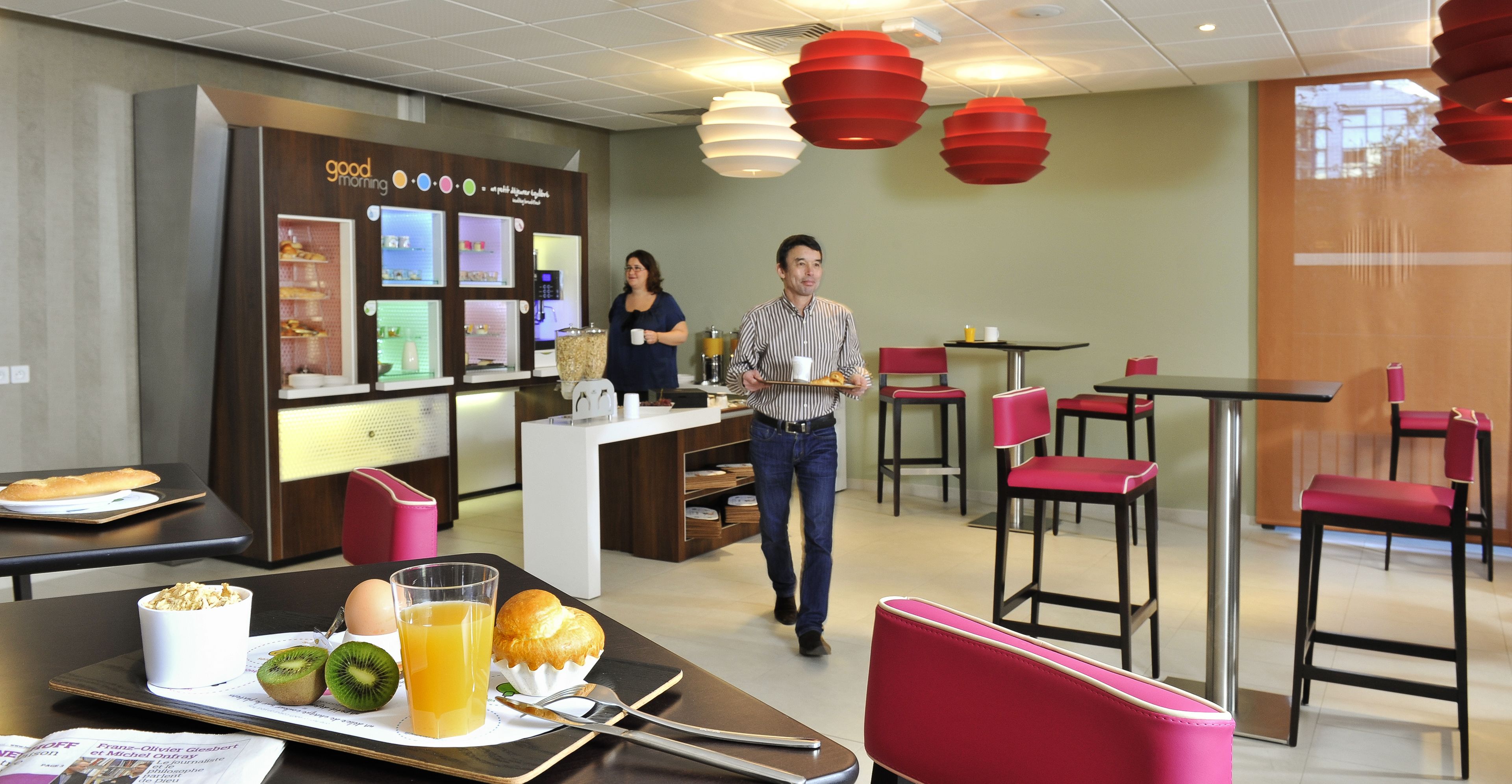 A modern breakfast area with a counter displaying food items, a seating area with red chairs, and a man walking through the space.