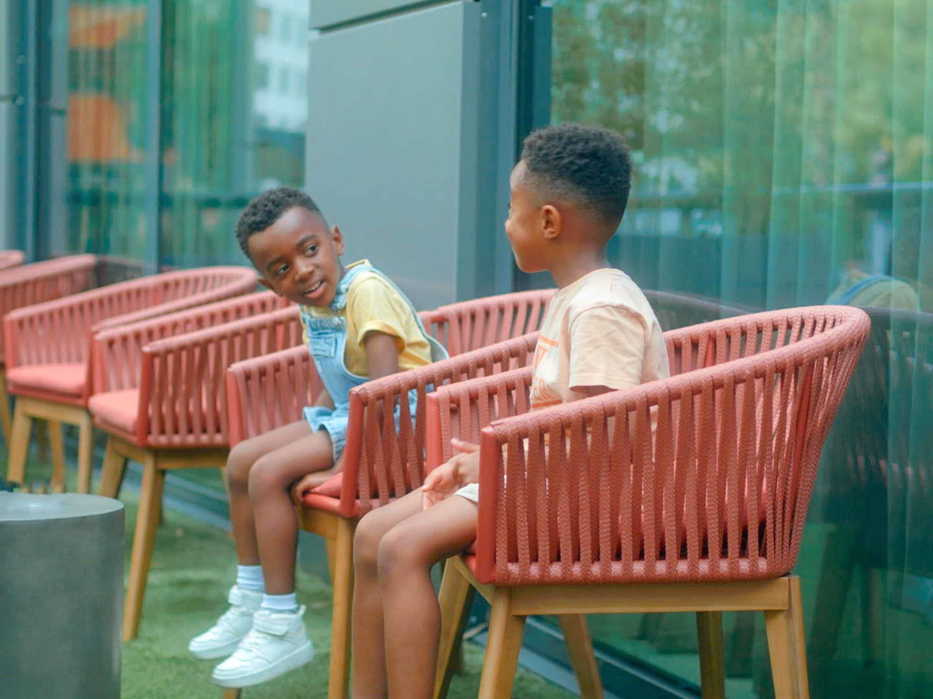Dos niños sentados en sillas de mimbre al aire libre, con un muro de cristal detrás.
