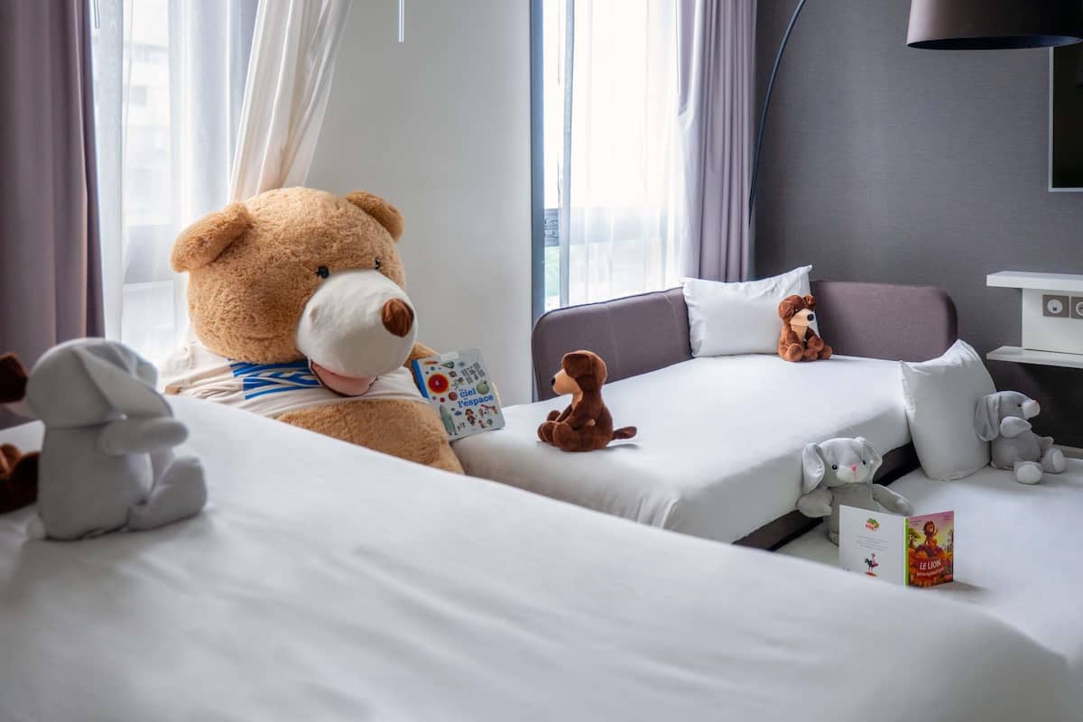 A hotel room with two beds, a large teddy bear on one bed, and a lamp on the wall.