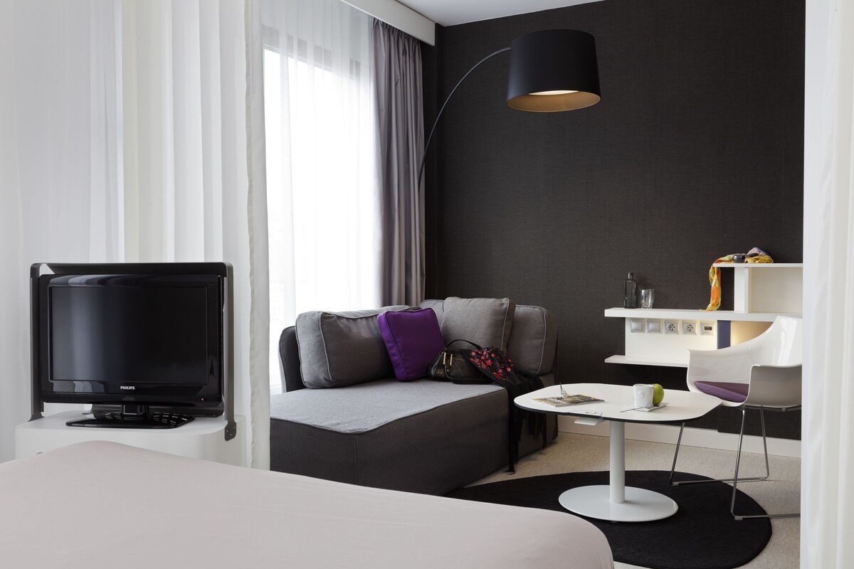 Een moderne hotelkamer met een flatscreen televisie, een bank met kussens, een klein rond tafeltje, een lamp en een witte console met decoratieve objecten.