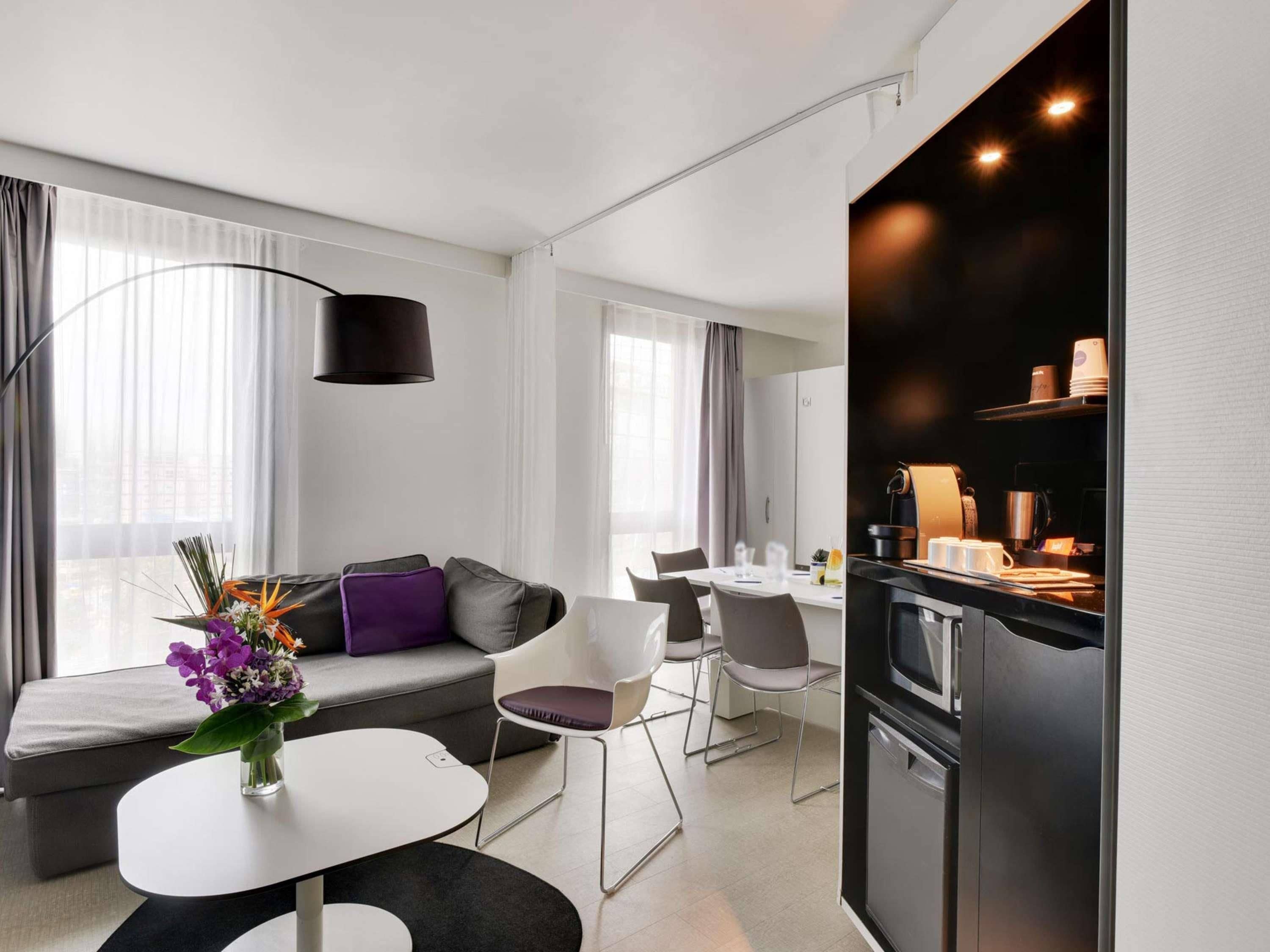 A modern hotel room with a dining area, kitchenette, and a sofa with a vase of flowers.