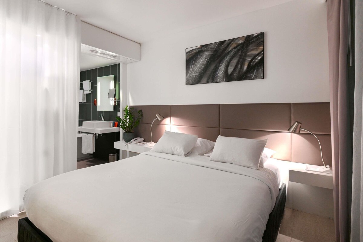 Une chambre d’hôtel moderne avec un grand lit, des tables de chevet, un miroir et une œuvre d’art accrochée au mur.
