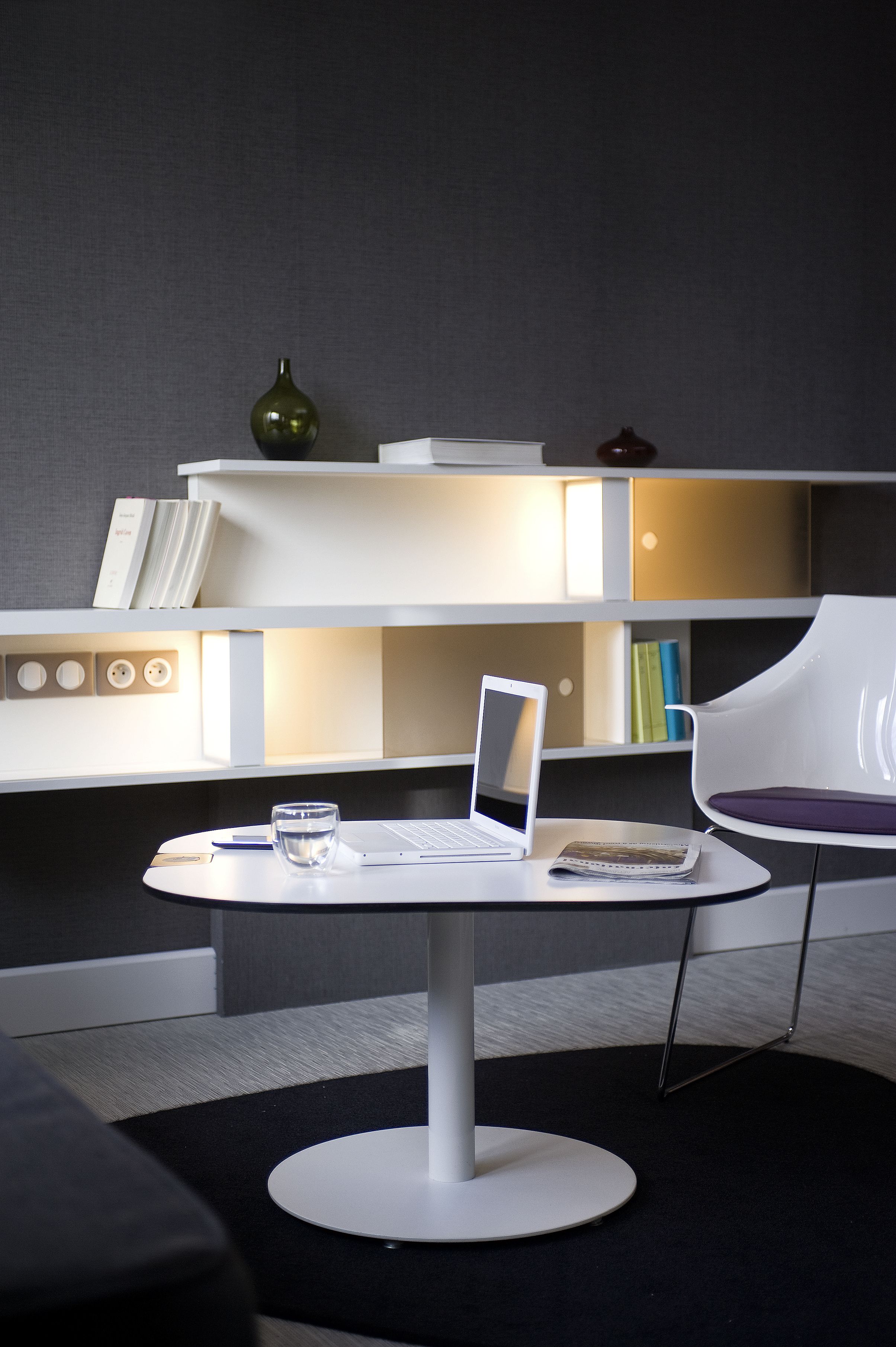 Een moderne kamer met een wit bureau, laptop en een glas water. Er staat een witte stoel en een boekenkast met boeken en decoratieve objecten.