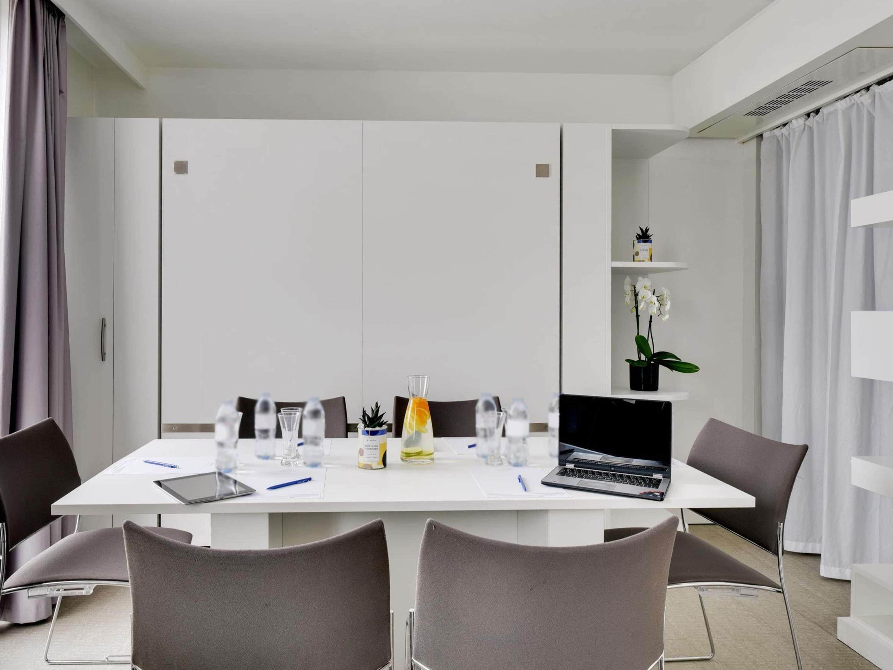 Una sala de reuniones moderna con una mesa blanca, sillas grises, una computadora portátil y una orquídea en maceta.