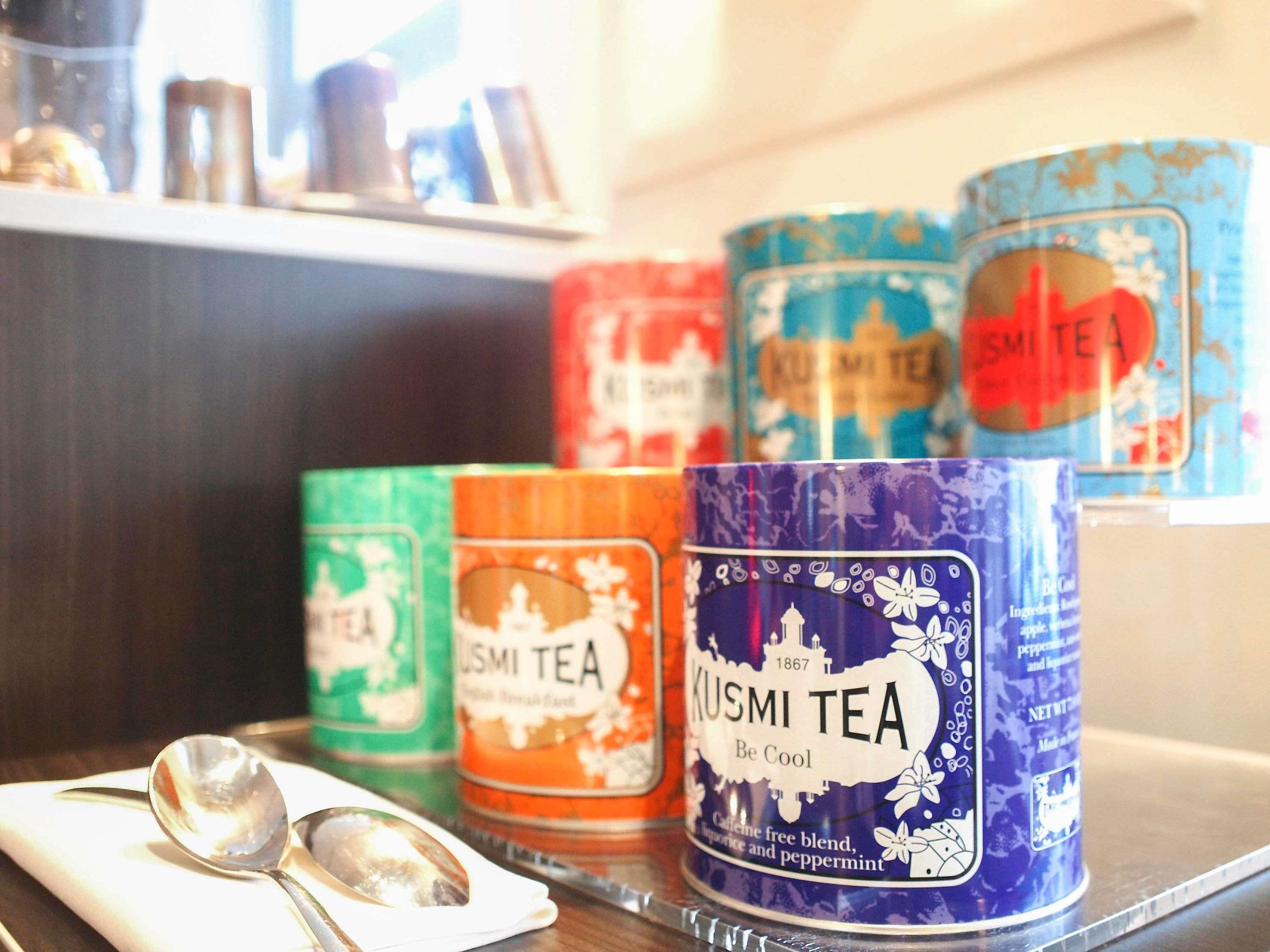 Een plank met verschillende theedozen, waaronder merken zoals LSMI TEA en KUSMI TEA, en een lepel op een servet.