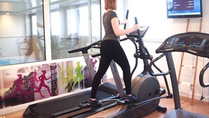 A woman using a treadmill in a gym with a large window, a television displaying workout information, and a poster of runners on the wall.