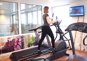 Une femme fait de l’exercice sur un tapis de course dans une salle de sport. On aperçoit une grande fenêtre, un téléviseur qui affiche des informations sur les exercices et une affiche représentant des coureurs sur le mur.