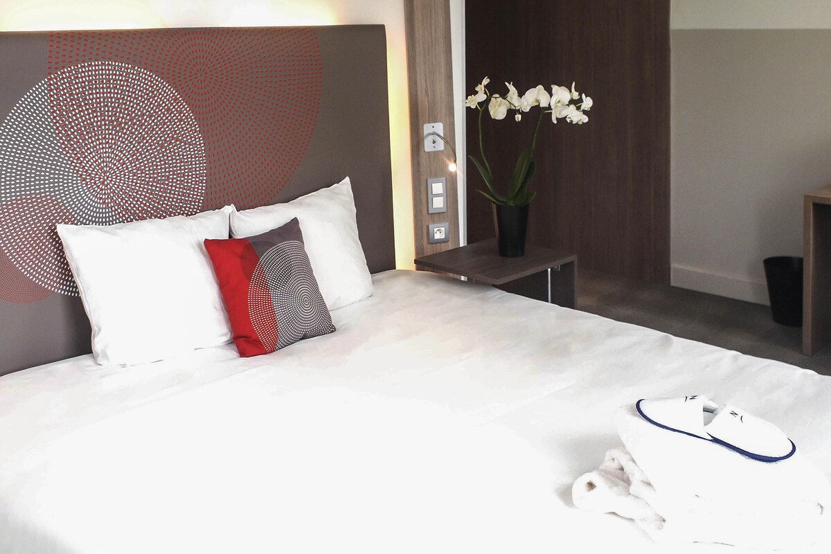 Una cama bien hecha con ropa de cama blanca, una almohada roja y negra, y un par de zapatillas blancas en el suelo.