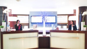 A hotel reception desk with a flat-screen TV, two receptionists, and decorative plants.
