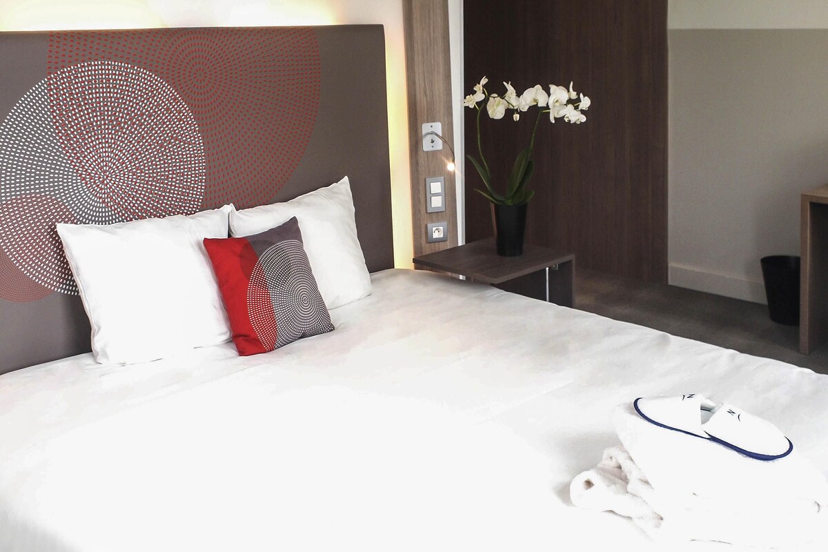 A neatly made bed with white bedding, a red and black pillow, and a pair of white slippers on the floor.