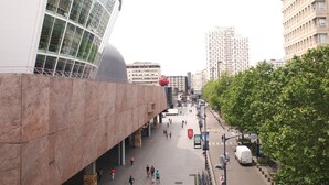 A modern urban square with a curved glass building, a red spherical sculpture, and a tree-lined street.