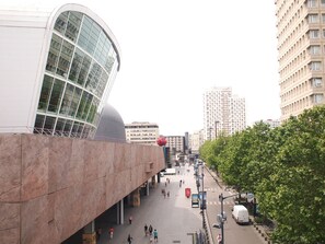 Une place urbaine moderne, avec un bâtiment aux façades courbes en verre, une sculpture sphérique rouge et une rue bordée d’arbres.
