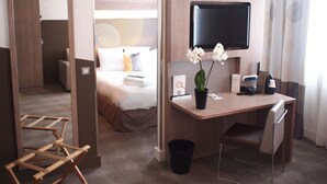 Habitación de hotel con cama, escritorio con silla, televisor, planta y cubo de basura.