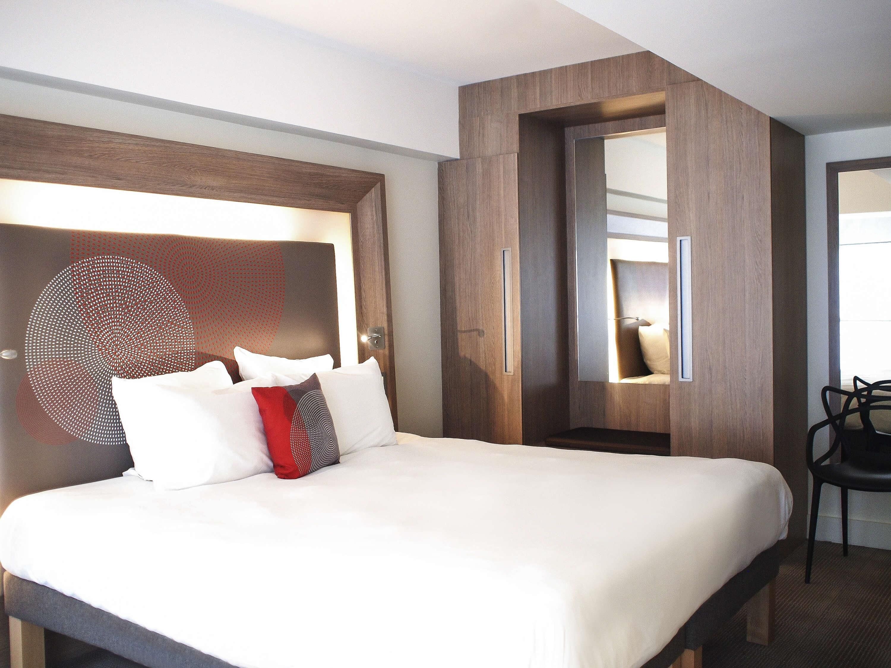 A hotel room with a large bed, a bedside table, a chair, and a mirror.