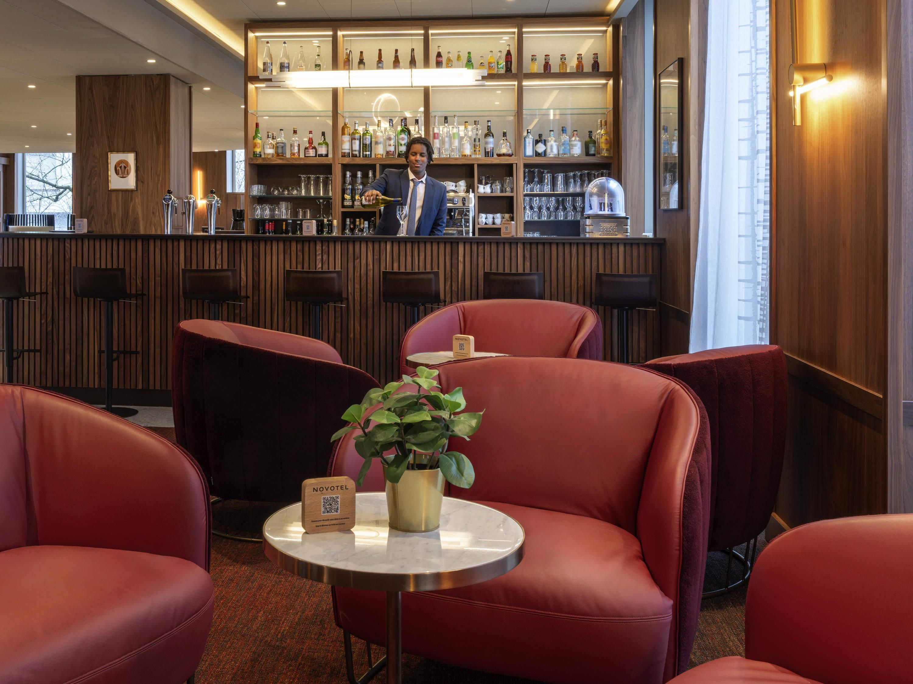 Una zona de bar con un cantinero, sillas tapizadas en rojo, una mesita con una planta y un letrero de Novotel.