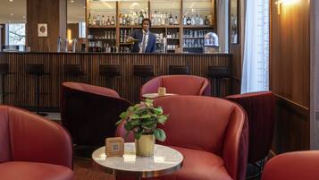 Una zona de bar con un cantinero, sillas tapizadas en rojo, una mesita con una planta y un letrero de Novotel.