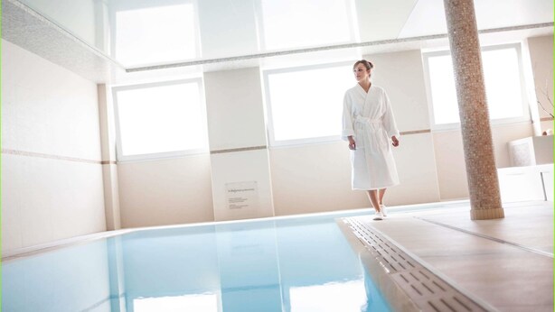 A person in a white bathrobe standing by a pool with a tiled floor and a pillar.