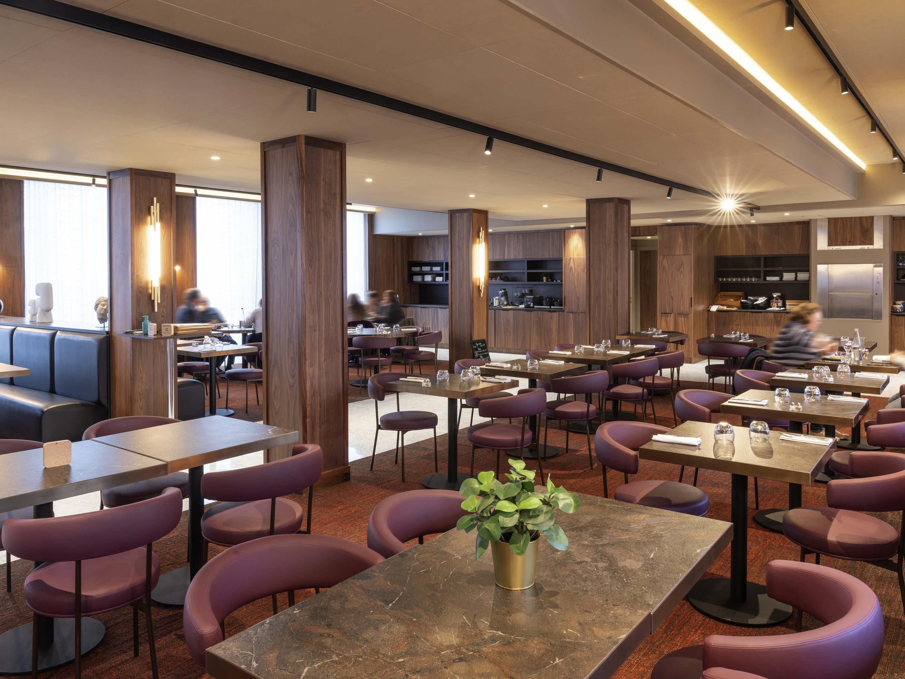 A modern restaurant with high tables and purple chairs, wooden accents, and a central plant.