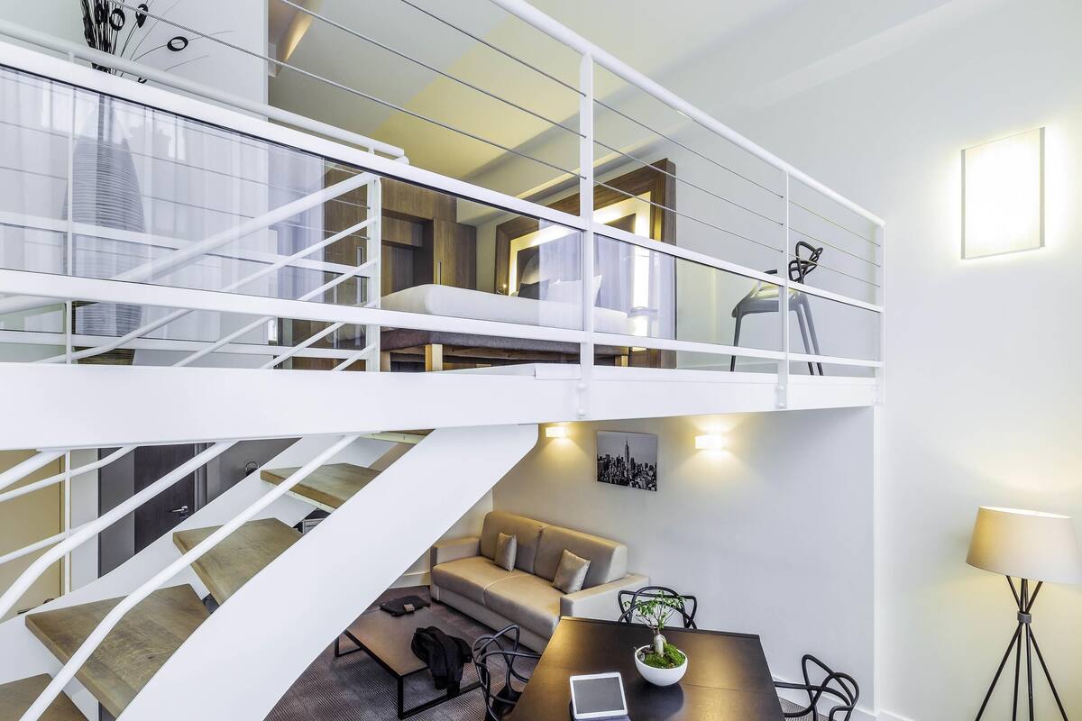 A modern interior with a staircase leading to a loft bedroom, a dining area with a table and chairs, and a small seating area with a sofa.