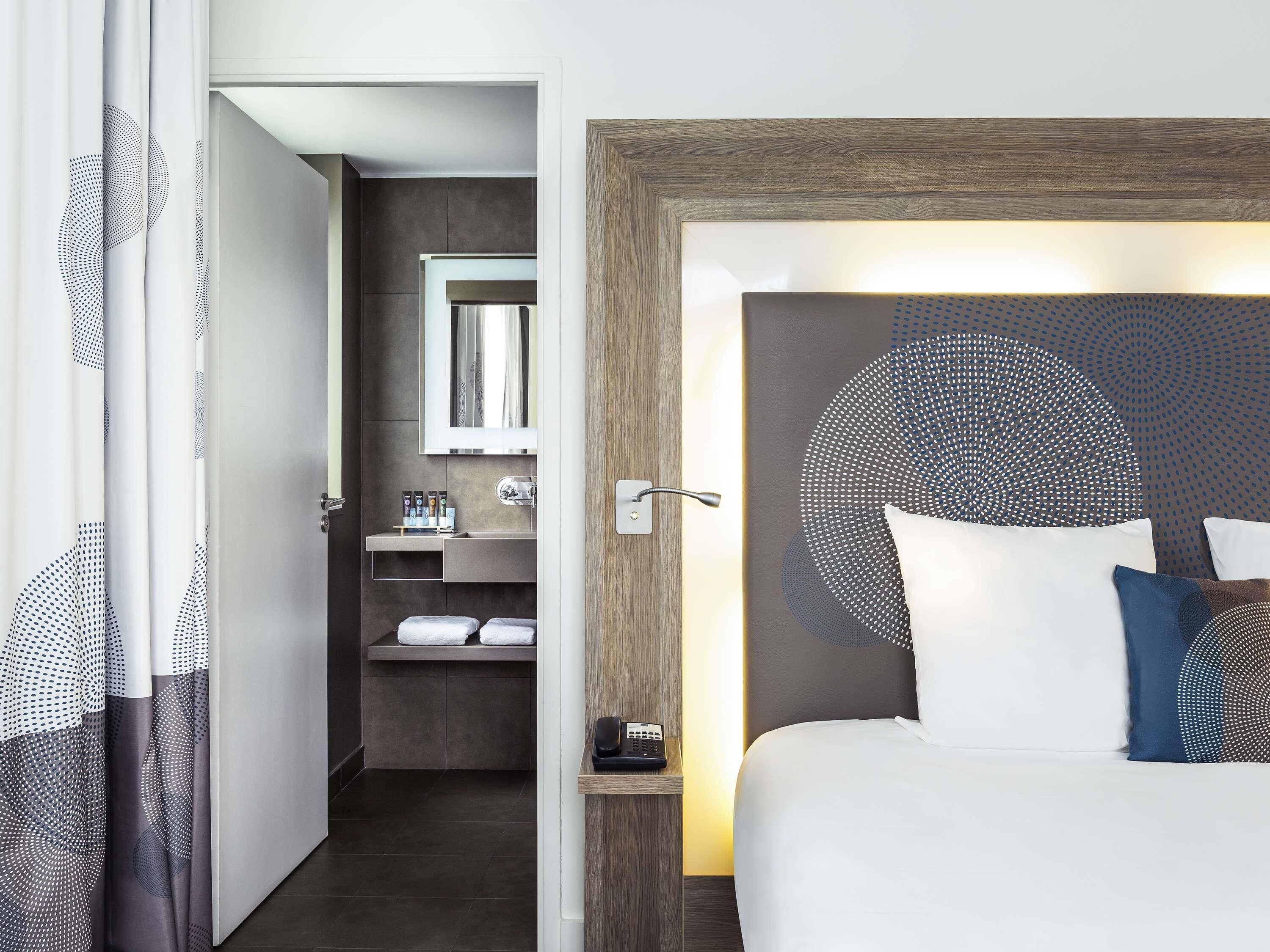 A hotel room with a bed, a bedside table with a phone, a mirror, a bathroom with a sink and towel rack.