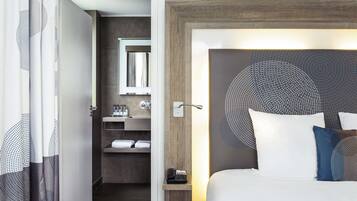 A hotel room with a bed, a bedside table with a phone, a mirror, a bathroom with a sink and towel rack.