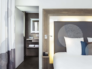 A hotel room with a bed, a bedside table with a phone, a mirror, a bathroom with a sink and towel rack.