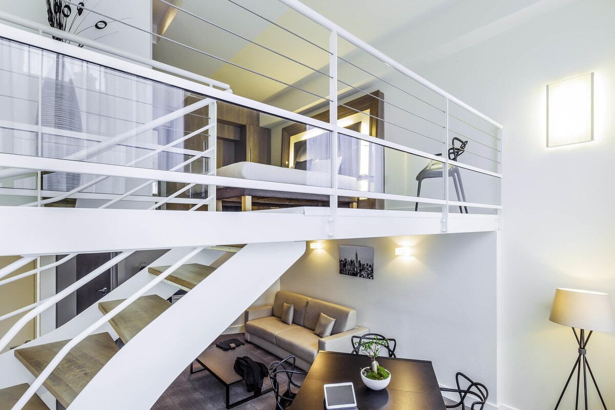 A modern interior with a staircase leading to a loft bedroom, a dining area with a table and chairs, and a small seating area with a sofa.