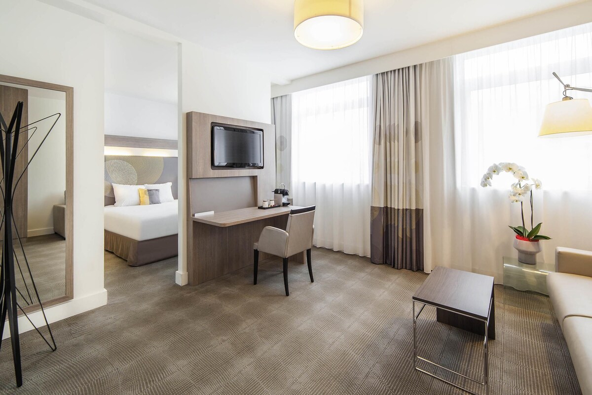 A hotel room with a bed, desk, chair, TV, and a potted plant.