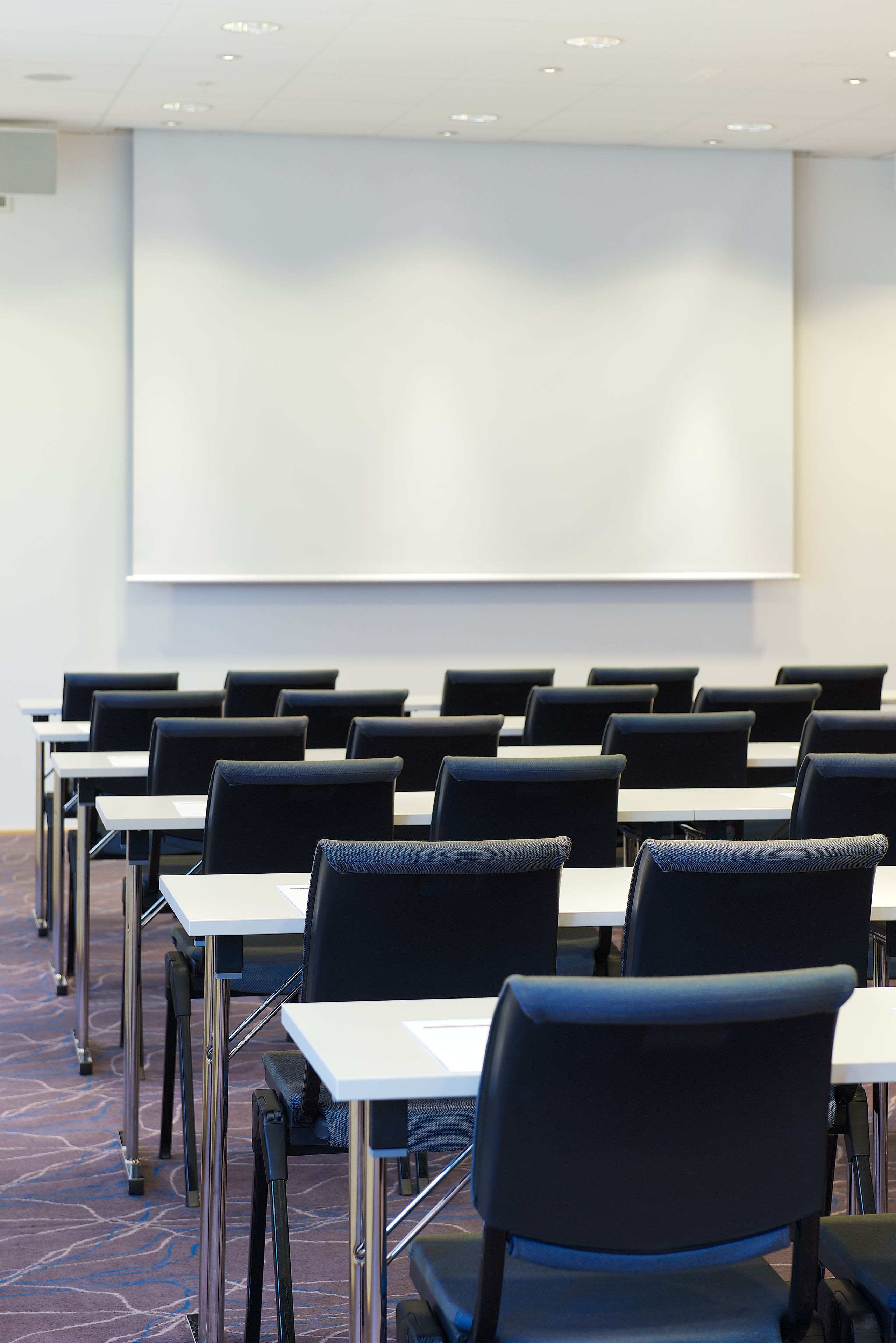 A conference room with rows of tables and chairs, a projector screen, and a carpeted floor.