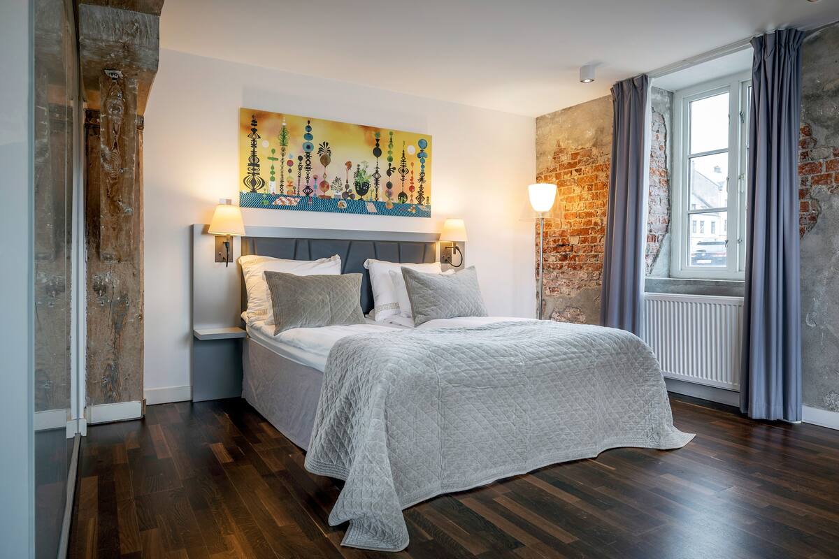 A bedroom with a bed, a headboard, a painting, a window with curtains, and a brick wall.