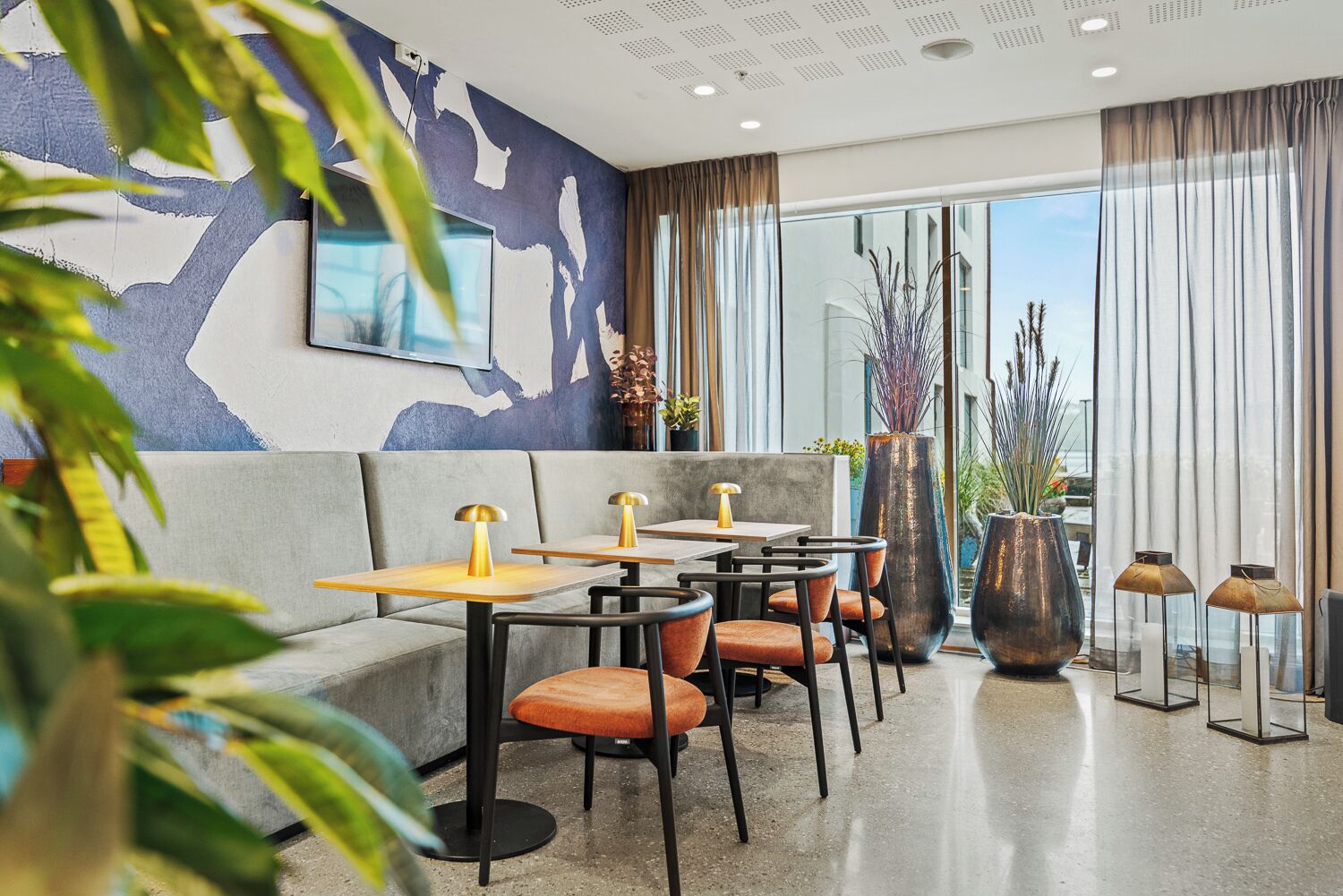 A modern hotel lobby with a seating area featuring sofas and tables, large windows with curtains, and a decorative wall mural.