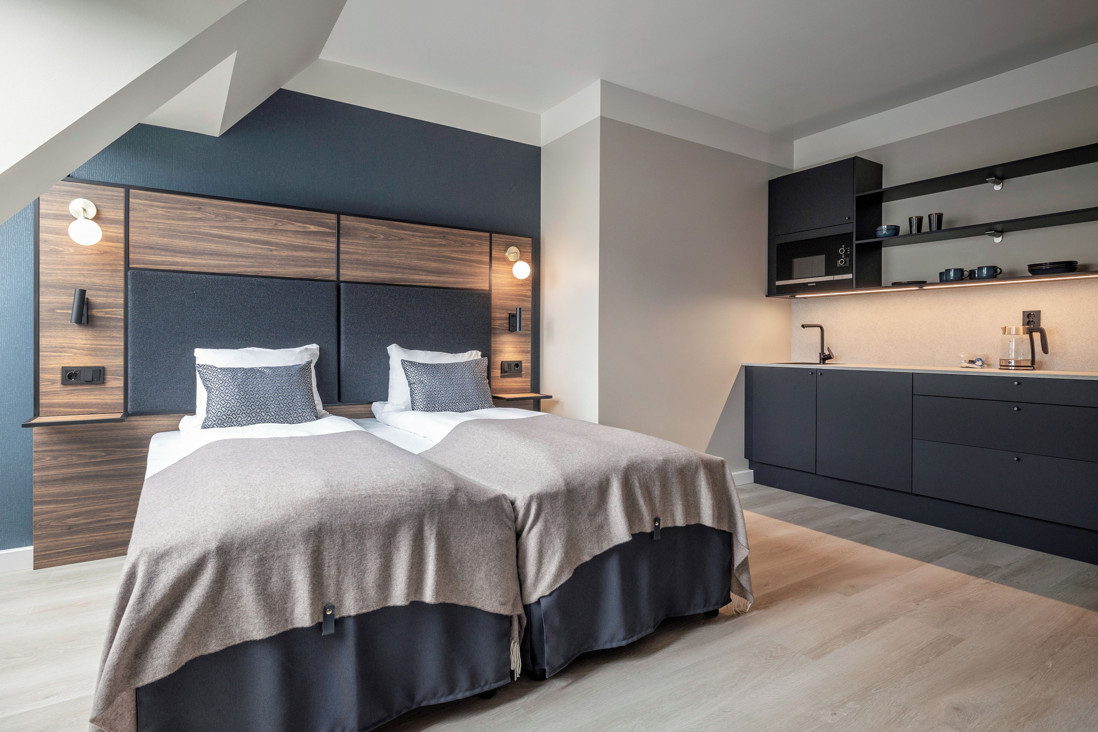 A modern bedroom with a large bed, built-in headboard, and a kitchenette area.