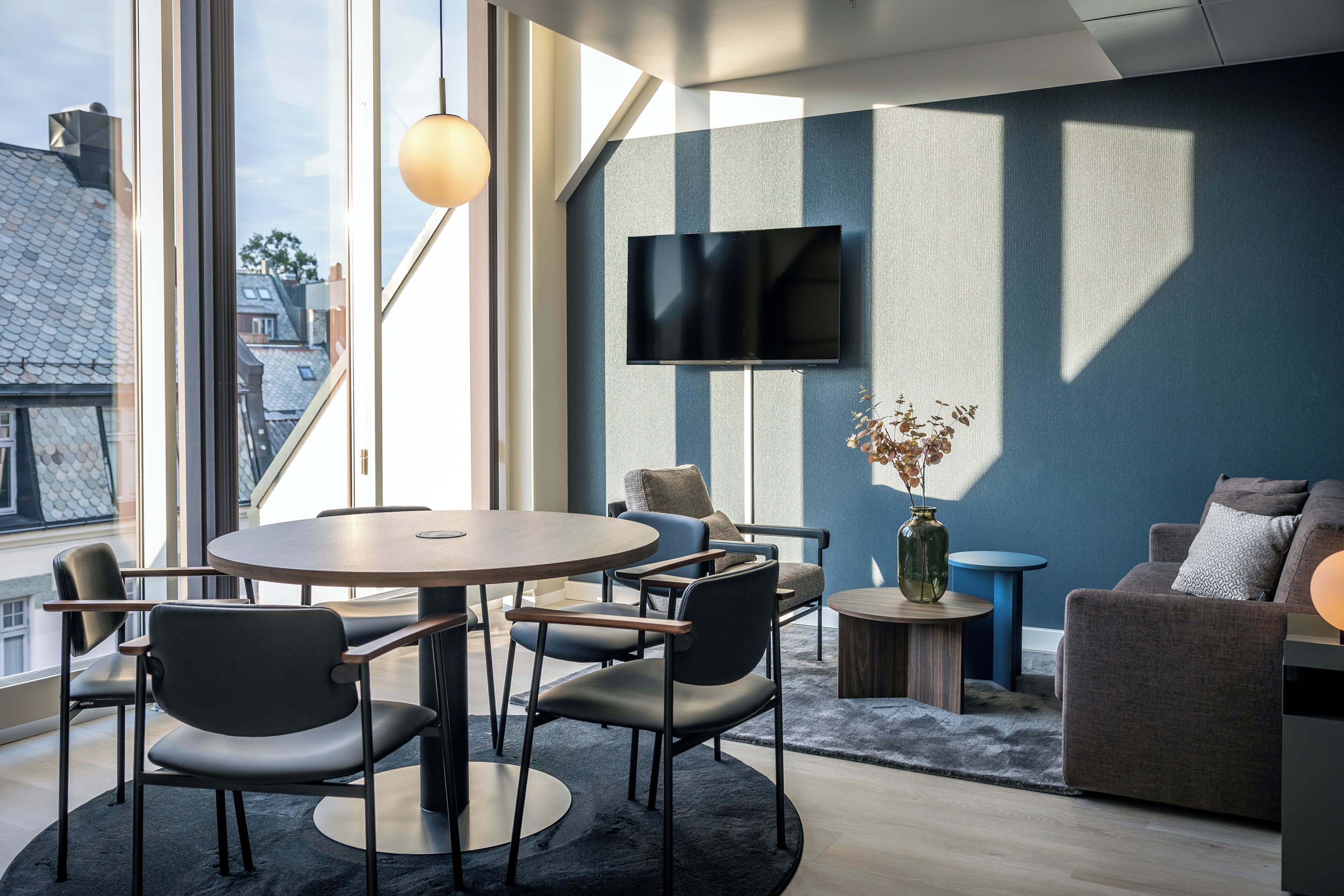 Ein modernes Wohnzimmer mit einem runden Tisch, Stühlen, einer Couch, einem an der Wand montierten Fernseher und Blick auf die Stadt durch große Fenster.