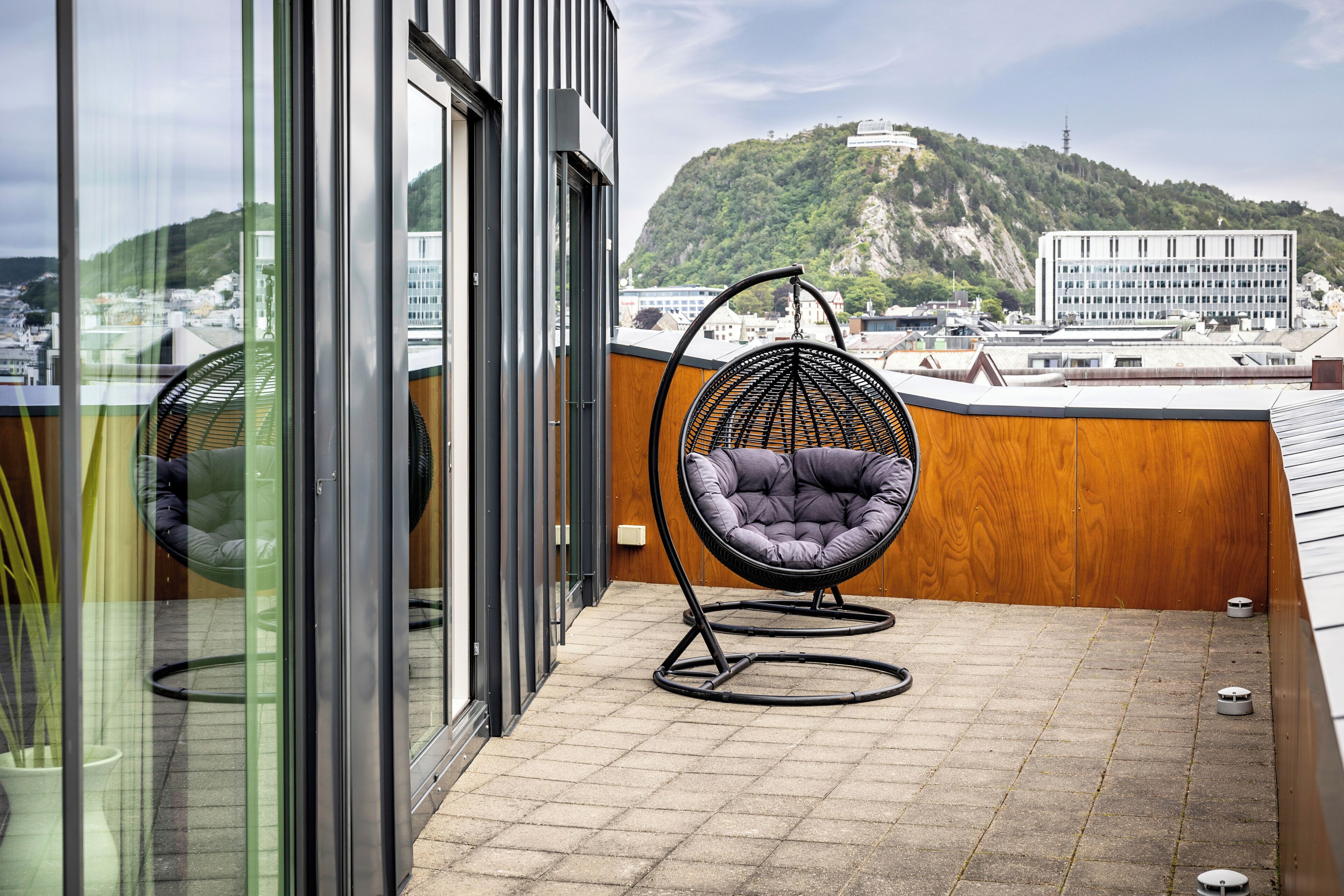A balcony with a hanging chair, a glass door, and a view of buildings and a hill.