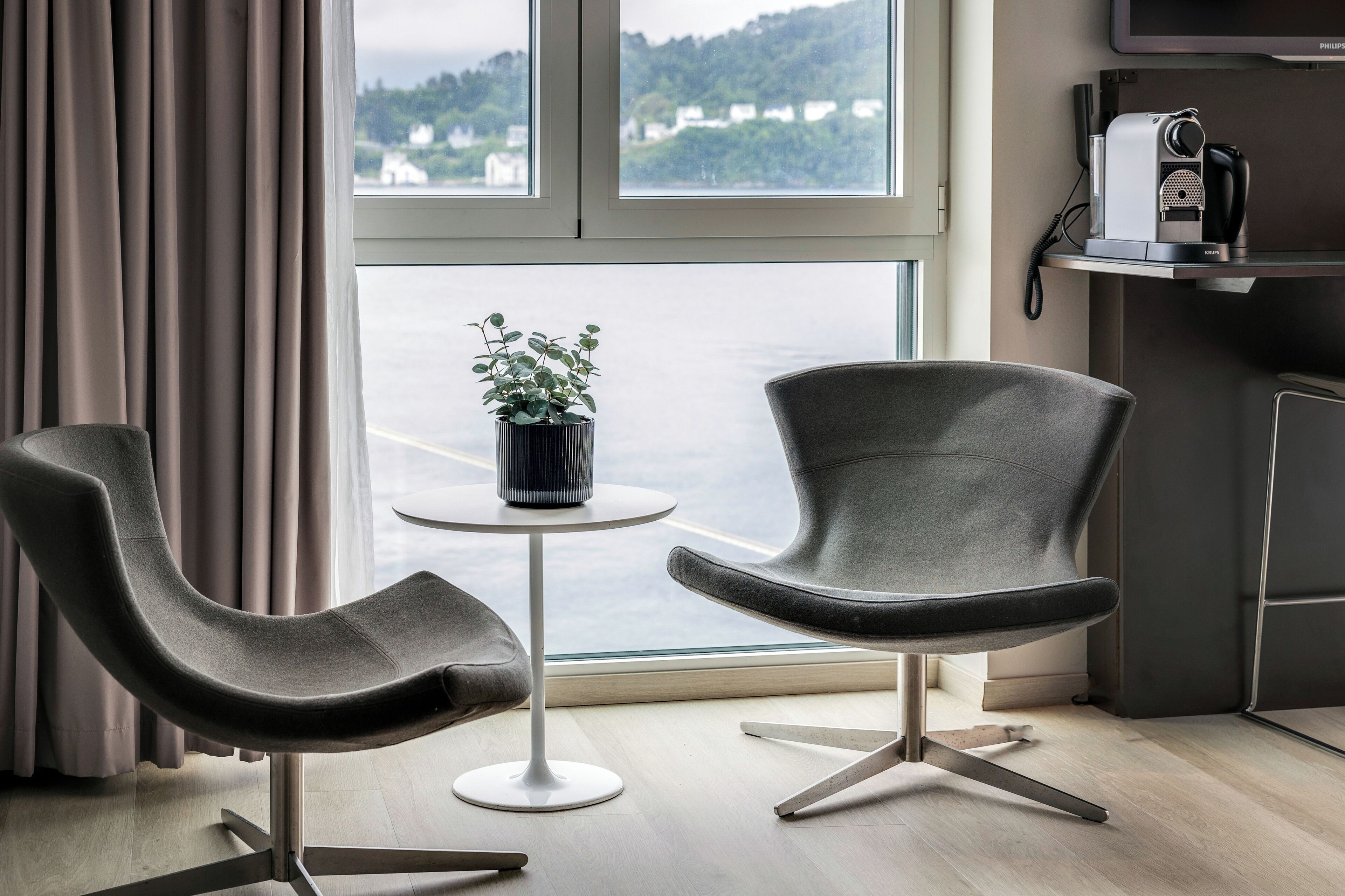 Habitación moderna con dos sillas, una mesita, una planta en maceta y vistas a una masa de agua a través de ventanas amplias.
