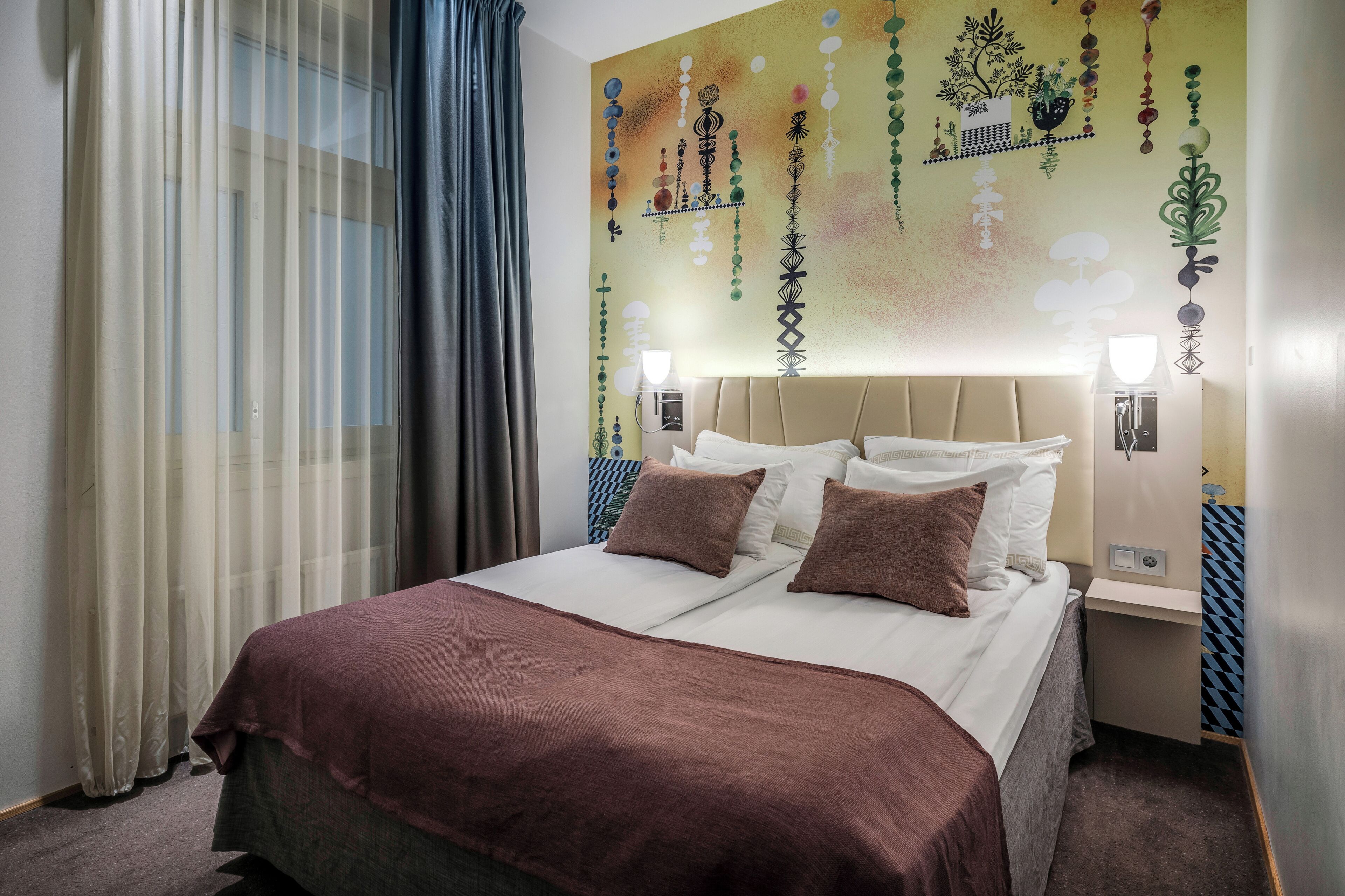 A hotel room with a bed, curtains, a wall with hanging decorations, and a nightstand with a lamp.