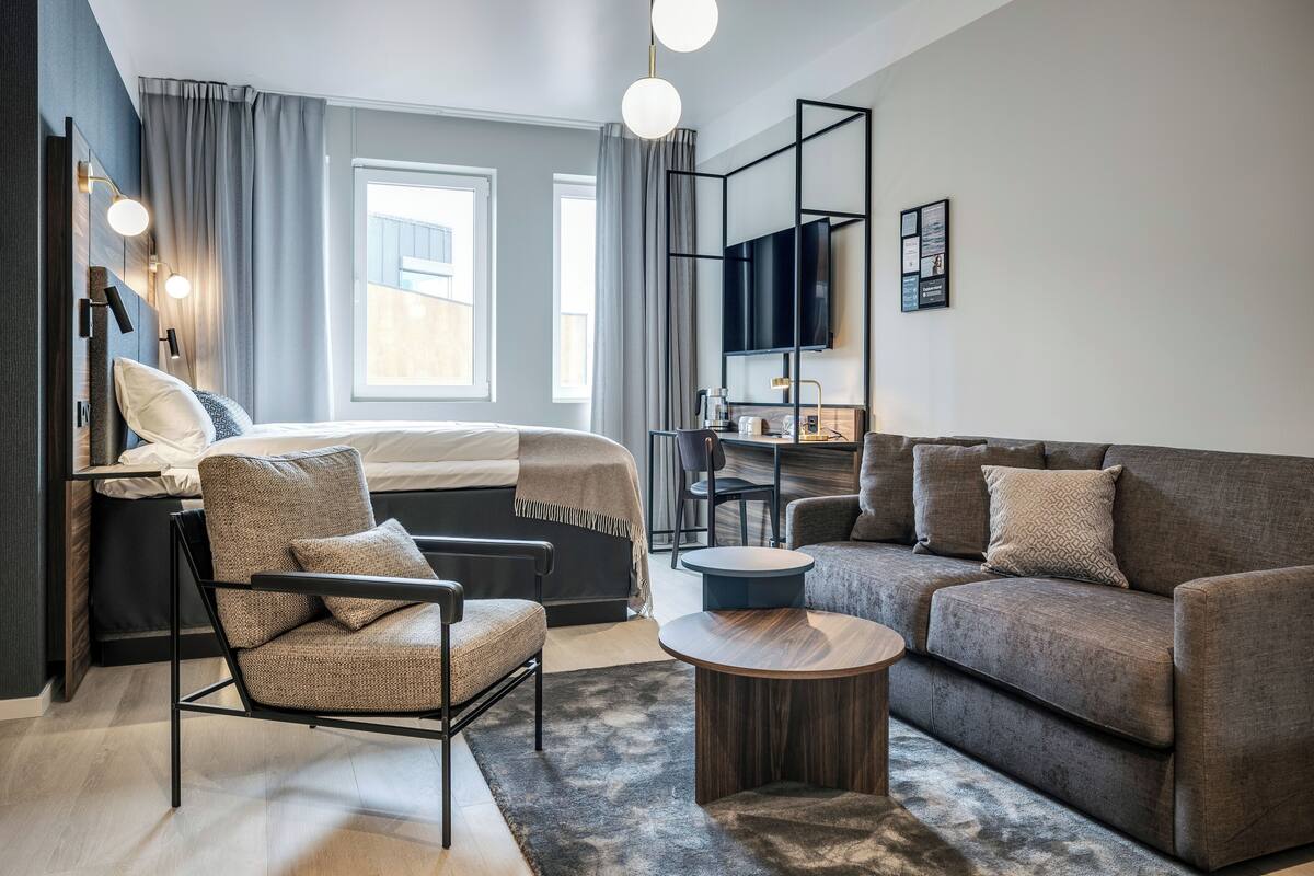 A modern hotel room with a bed, two sofas, a chair, a coffee table, a desk, and a TV.