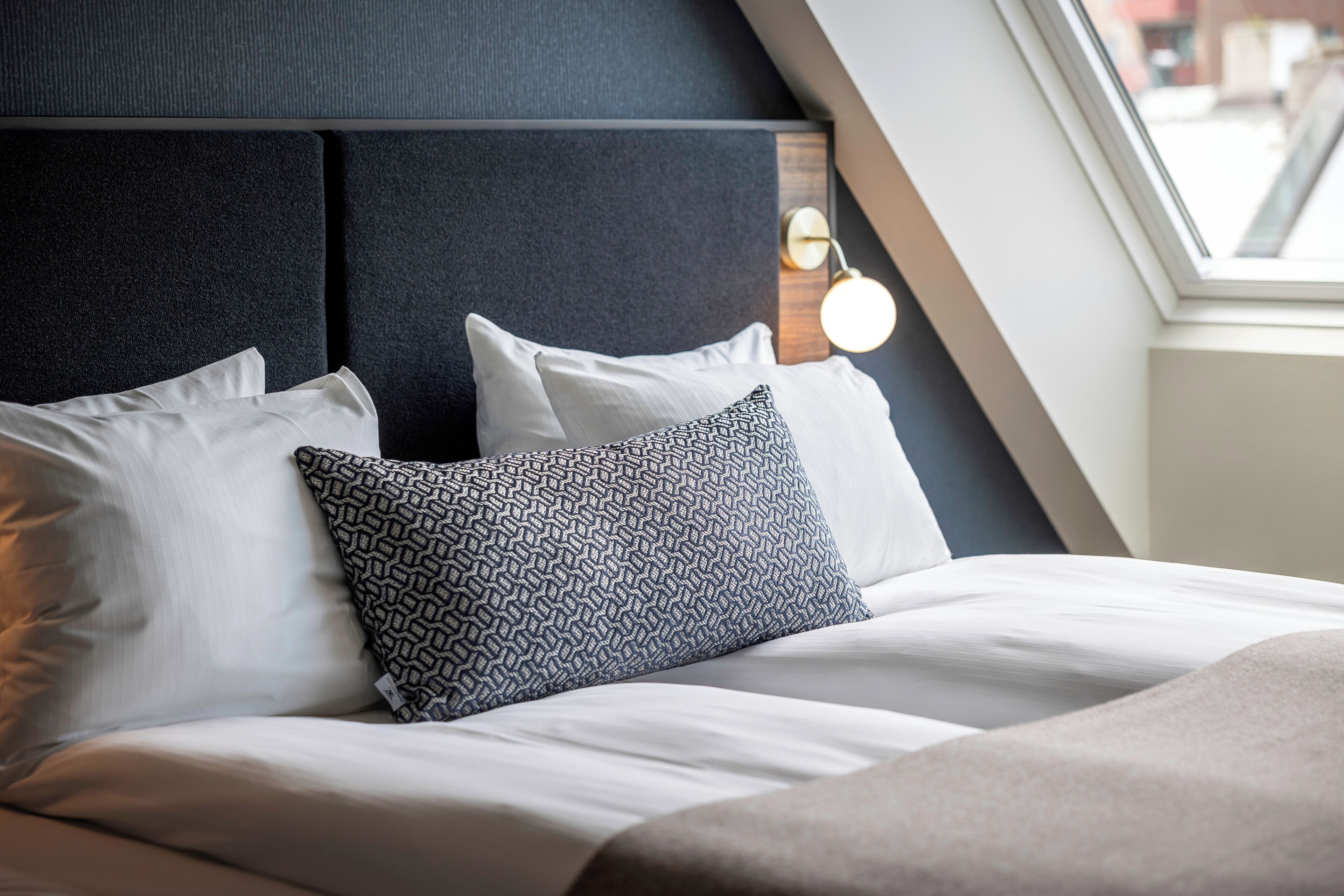 A neatly made bed with a patterned pillow, a headboard, and a window with a view of the outside.