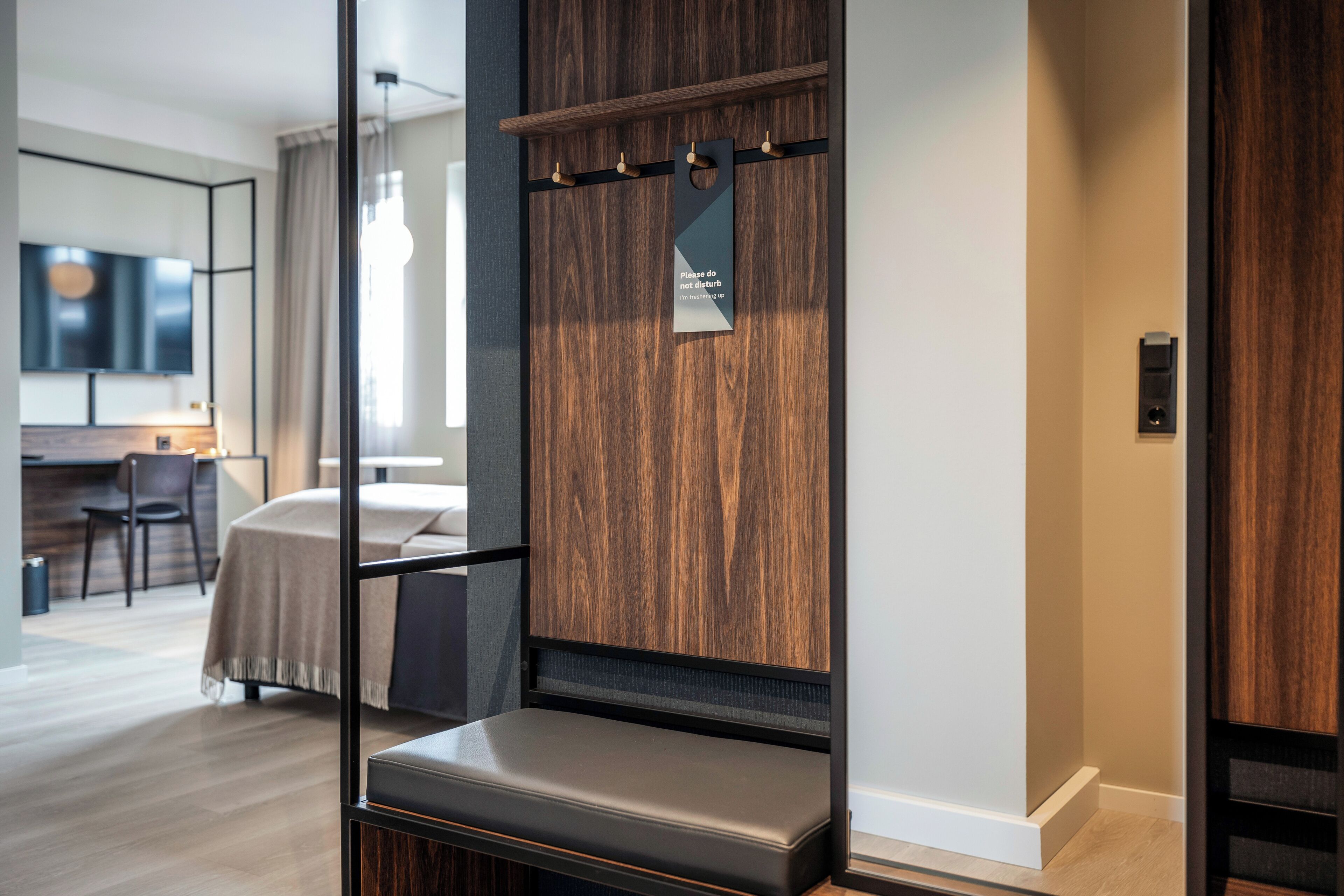 Une chambre d’hôtel moderne avec un mur d’accent en bois, un coin salon avec un banc, un téléviseur à écran plat, un bureau avec une chaise et un coin repas avec une table et des chaises.