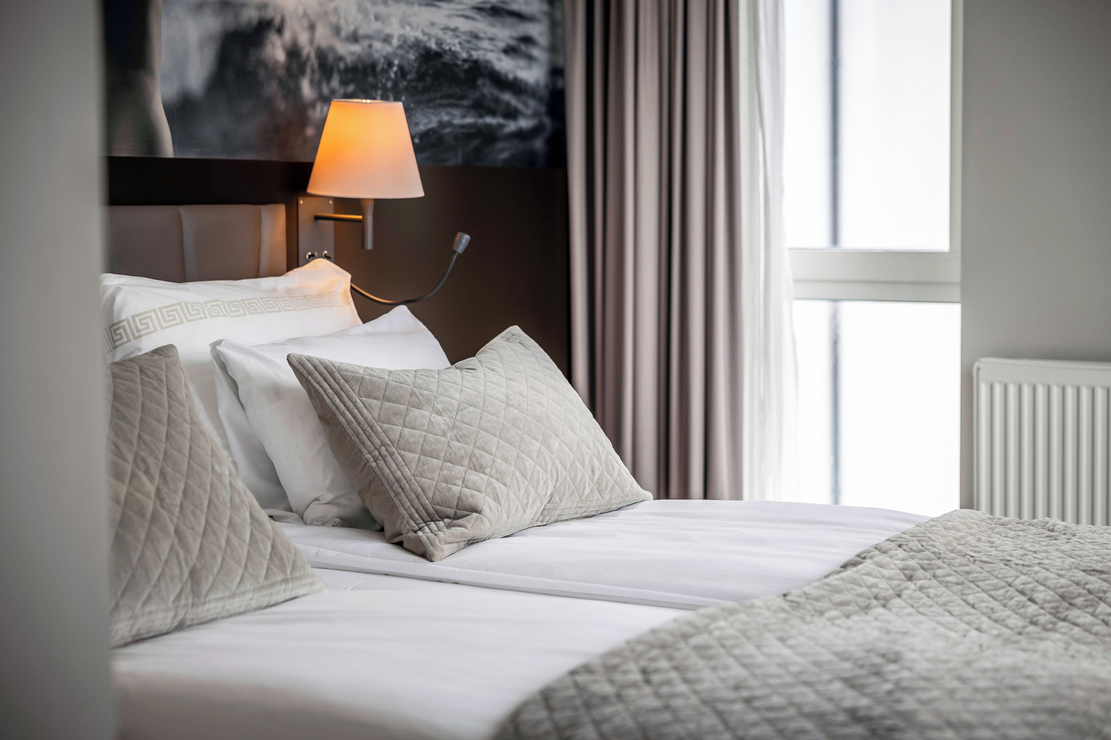 A neatly made bed with a quilted blanket and pillows, a bedside lamp, and a view of a window with curtains.