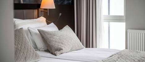 Una cama bien hecha con una colcha acolchada y almohadas, una lámpara de noche y vistas a una ventana con cortinas.