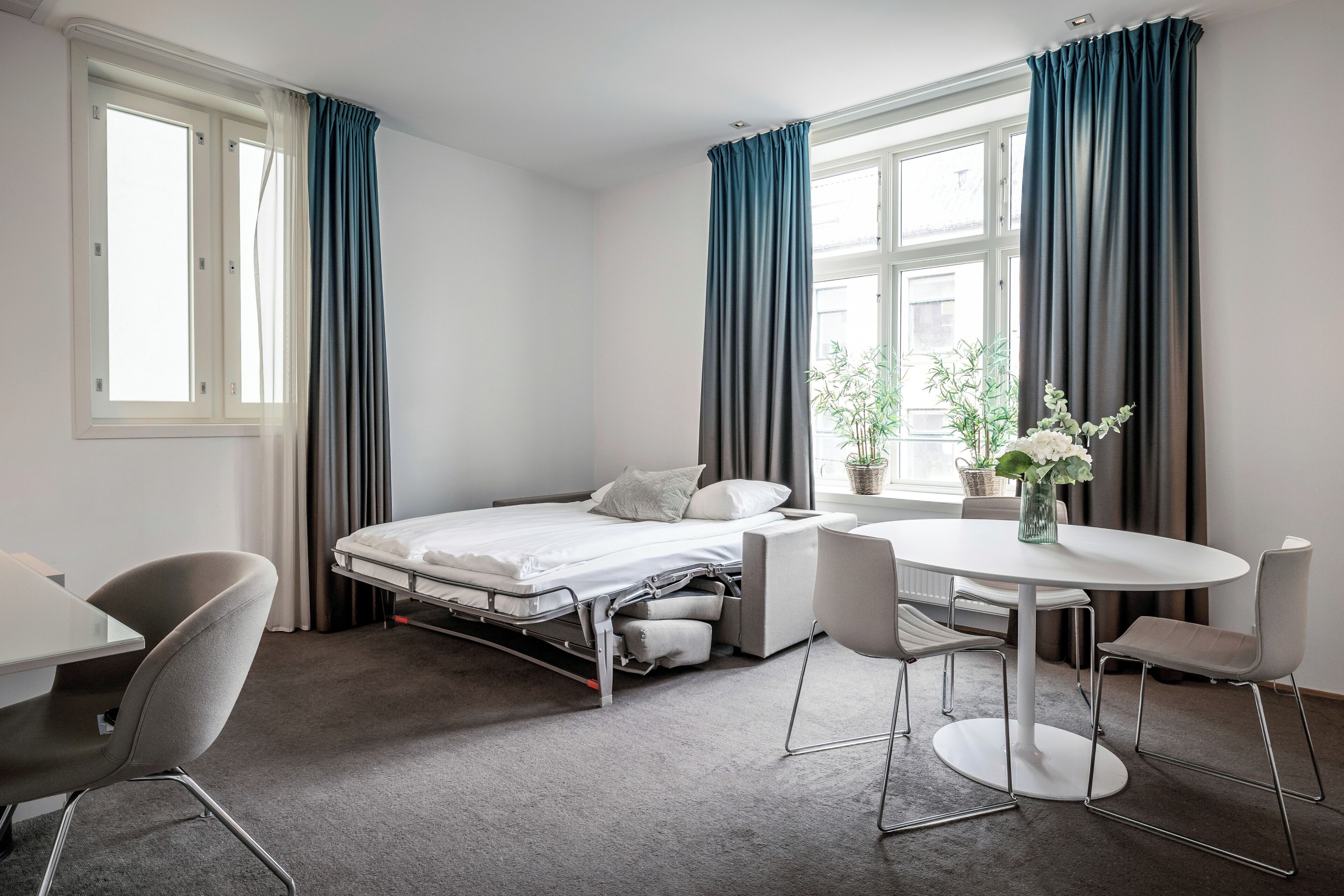 A compact hotel room with a bed, a small dining table, a chair, and a potted plant.