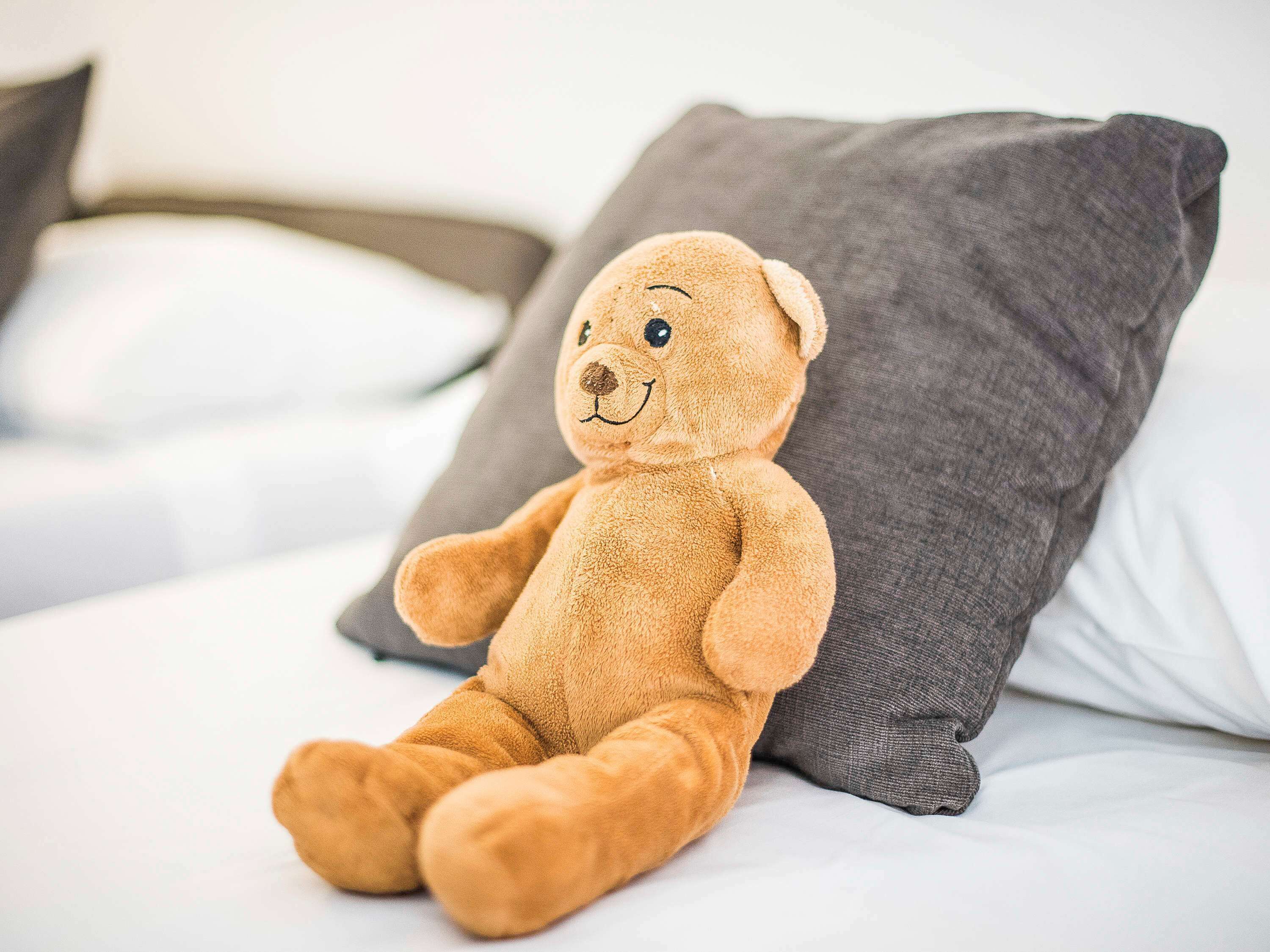 Um urso de pelúcia aconchegante sentado sobre uma cama com lençóis brancos e almofadas cinzentas.