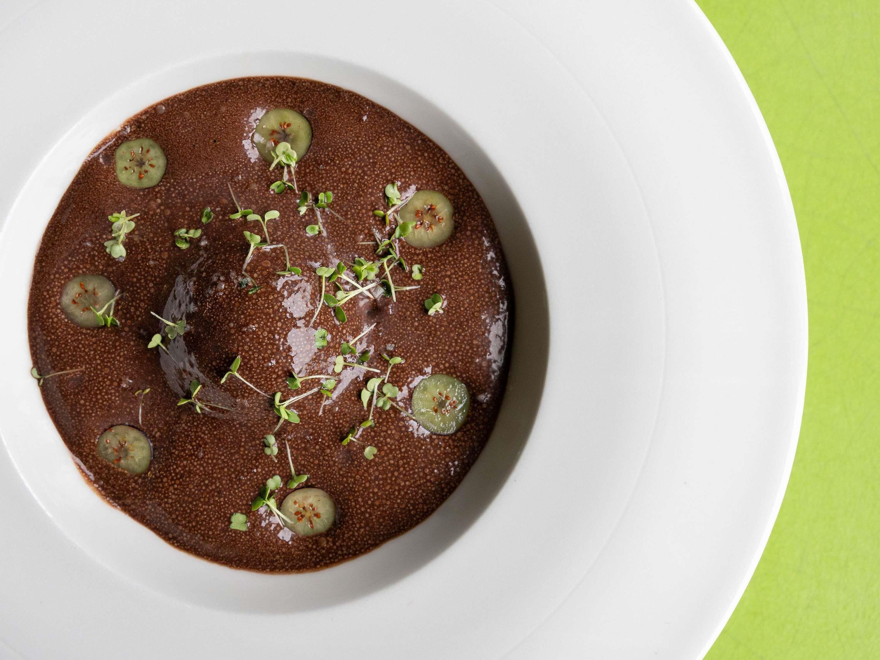 Uma tigela de sopa escura decorada com microverdes e fatias de pepino sobre um prato branco.