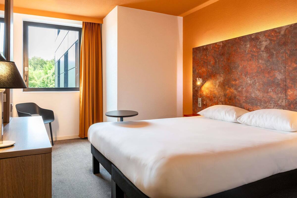Une chambre d’hôtel avec un lit, un bureau, une chaise, une table ronde et une fenêtre avec des rideaux.