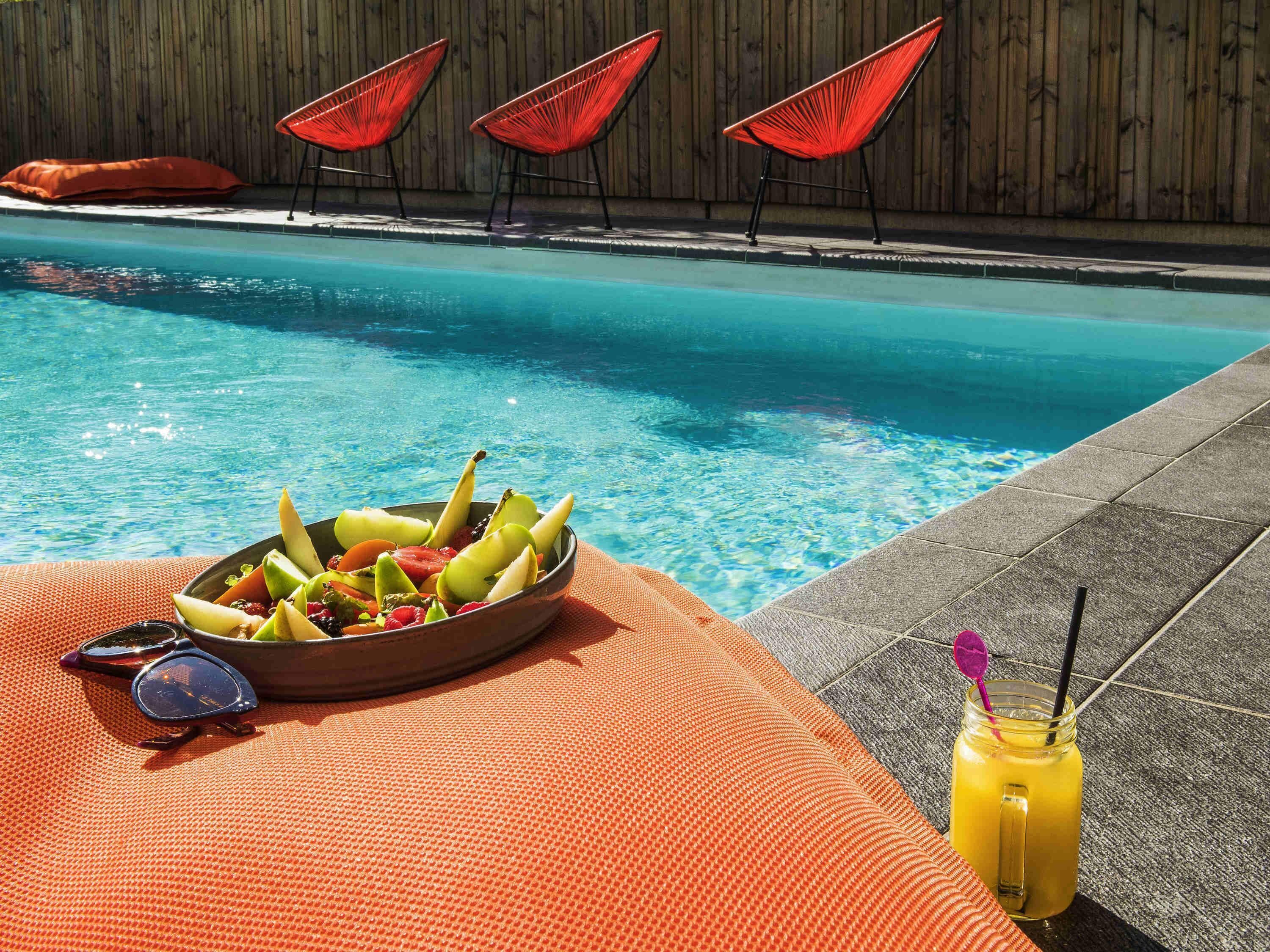 Une scène de bord de piscine avec un saladier de fruits, des lunettes de soleil et une boisson.