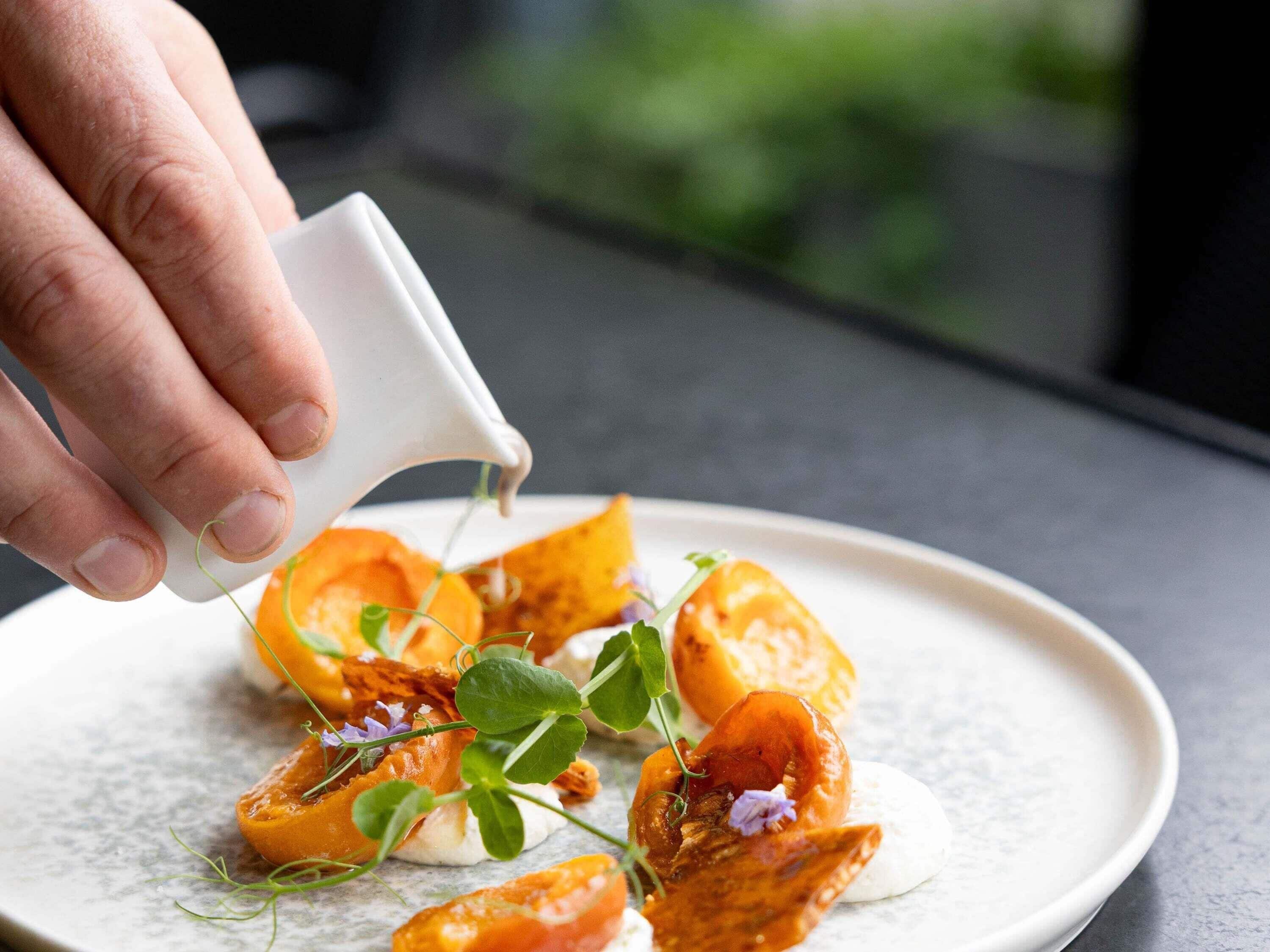 A hand pouring a liquid onto a plate with food and garnishes.
