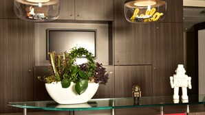 Een modern receptiebalie met een glazen blad, een witte plantenbak met diverse planten, en twee hanglampen met een 'hello'-bordje.