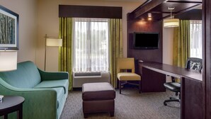 Habitación de hotel con sof á verde, un escritorio, una silla, un reposapiés, un televisor de pantalla plana y un ventanal con cortinas.