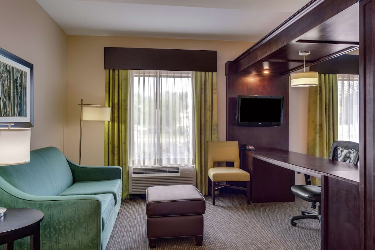 A hotel room with a green sofa, a desk, a chair, an ottoman, a flat-screen TV, and a large window with curtains.